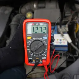 Ręka w czarnej rękawiczce trzyma multimetr, pokazując 12.72V. To pokazuje, jak zmierzyć baterię za pomocą multimetru.