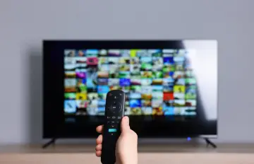 Dłoń trzyma pilot, celując w ekran telewizora z mozaiką obrazów. Szukasz częstotliwości kanału TVN, by obejrzeć ulubiony serial.