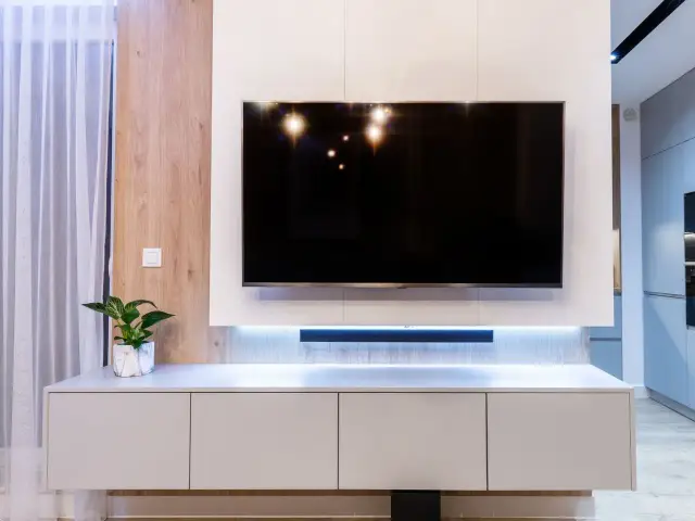 Nowoczesny salon z telewizorem na ścianie. Pod nim biała szafka RTV, a za nią subtelne podświetlenie. W rogu stoi roślina w doniczce.